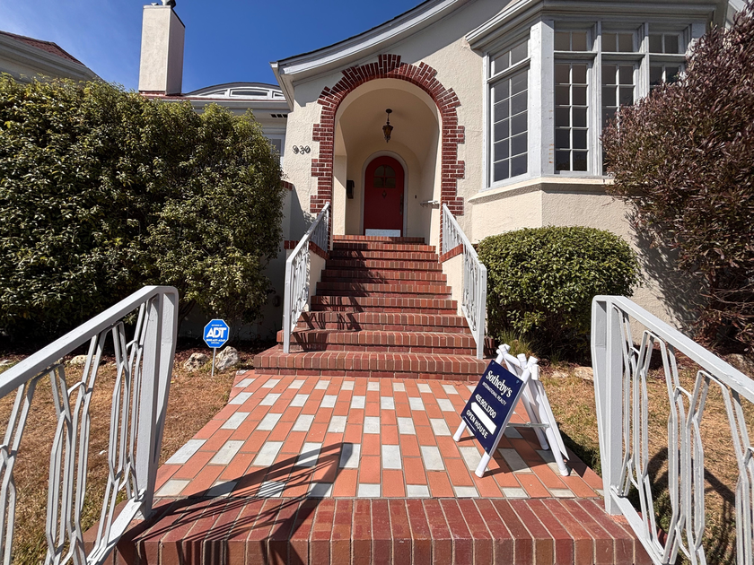 A brick staircase with white railings leads to a red door under an arch with a hanging light, flanked by bushes and a Sotheby’s real estate sign.