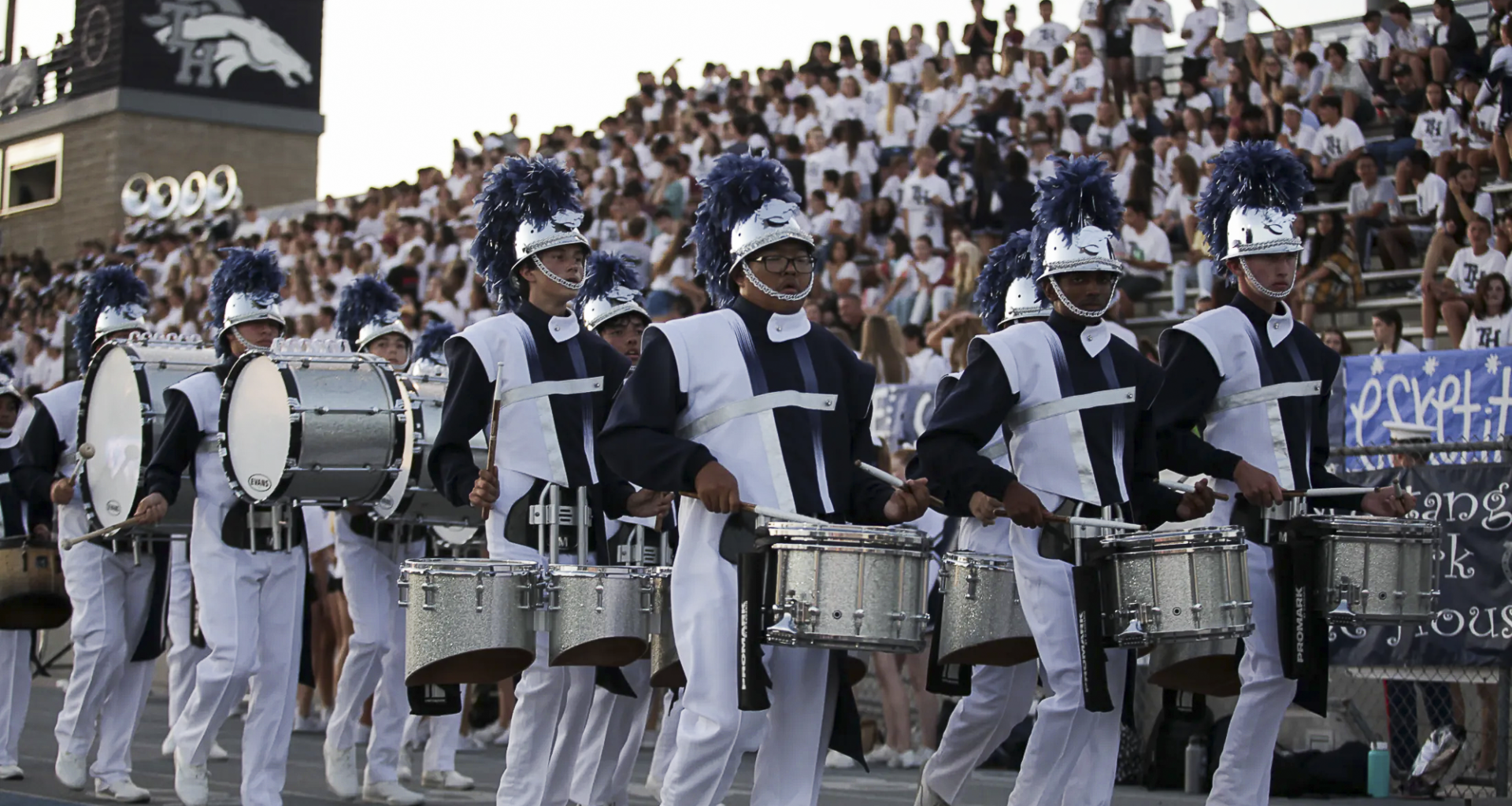 Students in Trabuco Hills High School’s marching band drumline, wearing navy and white uniforms with plumed helmets, perform during a football game in front of a packed stadium crowd.
