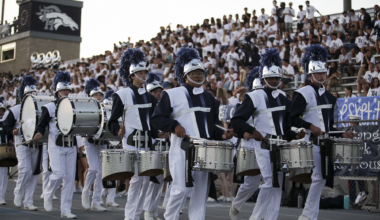 Students in Trabuco Hills High School’s marching band drumline, wearing navy and white uniforms with plumed helmets, perform during a football game in front of a packed stadium crowd.