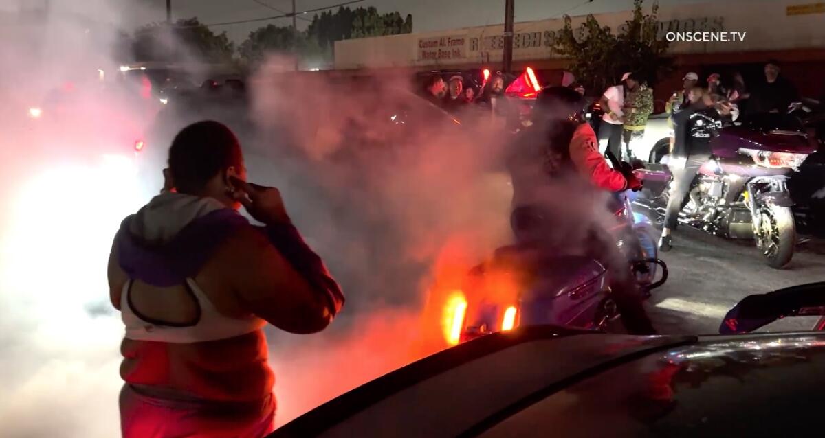 A motorcycle burns rubber as a man in the foreground plugs his ears.