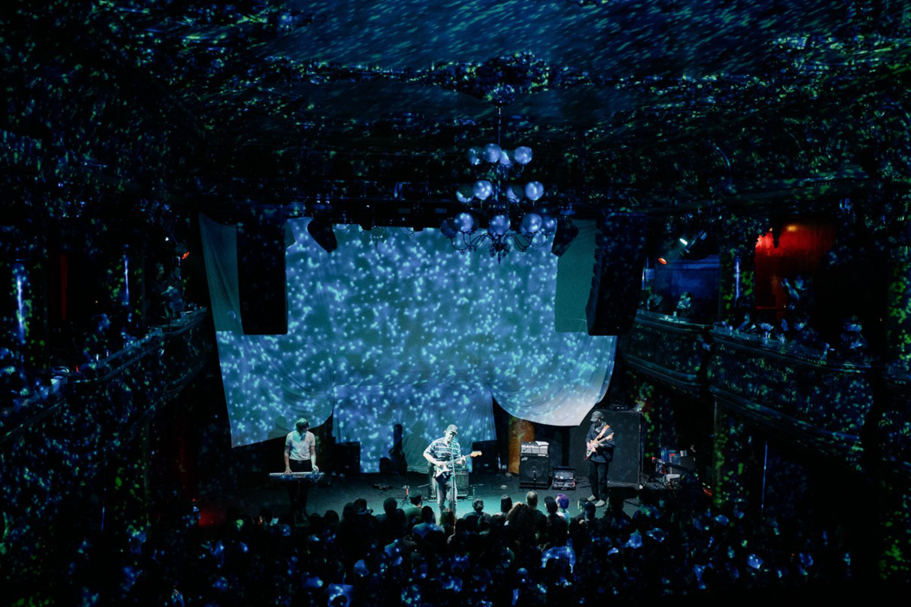 A band performs on stage with blue star-like light projections creating a cosmic atmosphere, while an audience watches in a dark, ornate venue.