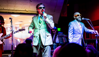 Two men in suits and sunglasses sing on stage with microphones, accompanied by a guitarist and drummer in a colorful, lively setting.