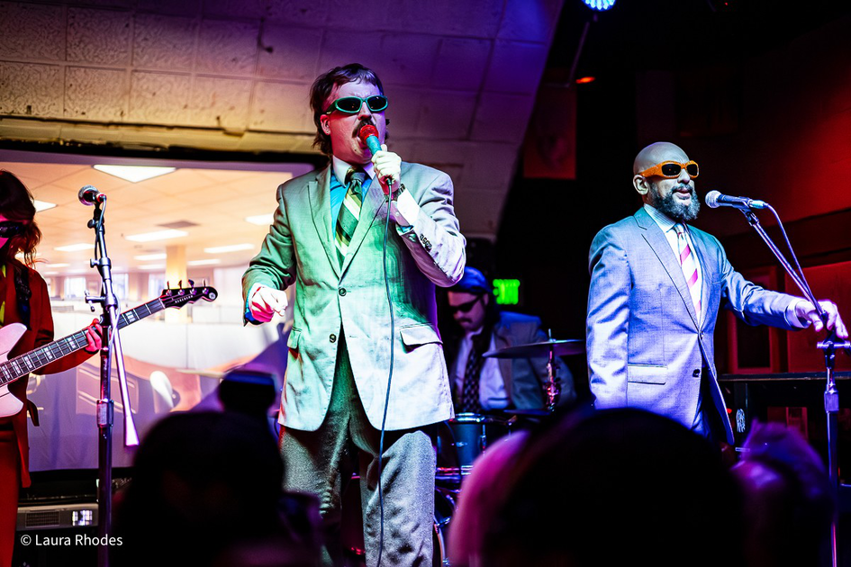Two men in suits and sunglasses sing on stage with microphones, accompanied by a guitarist and drummer in a colorful, lively setting.