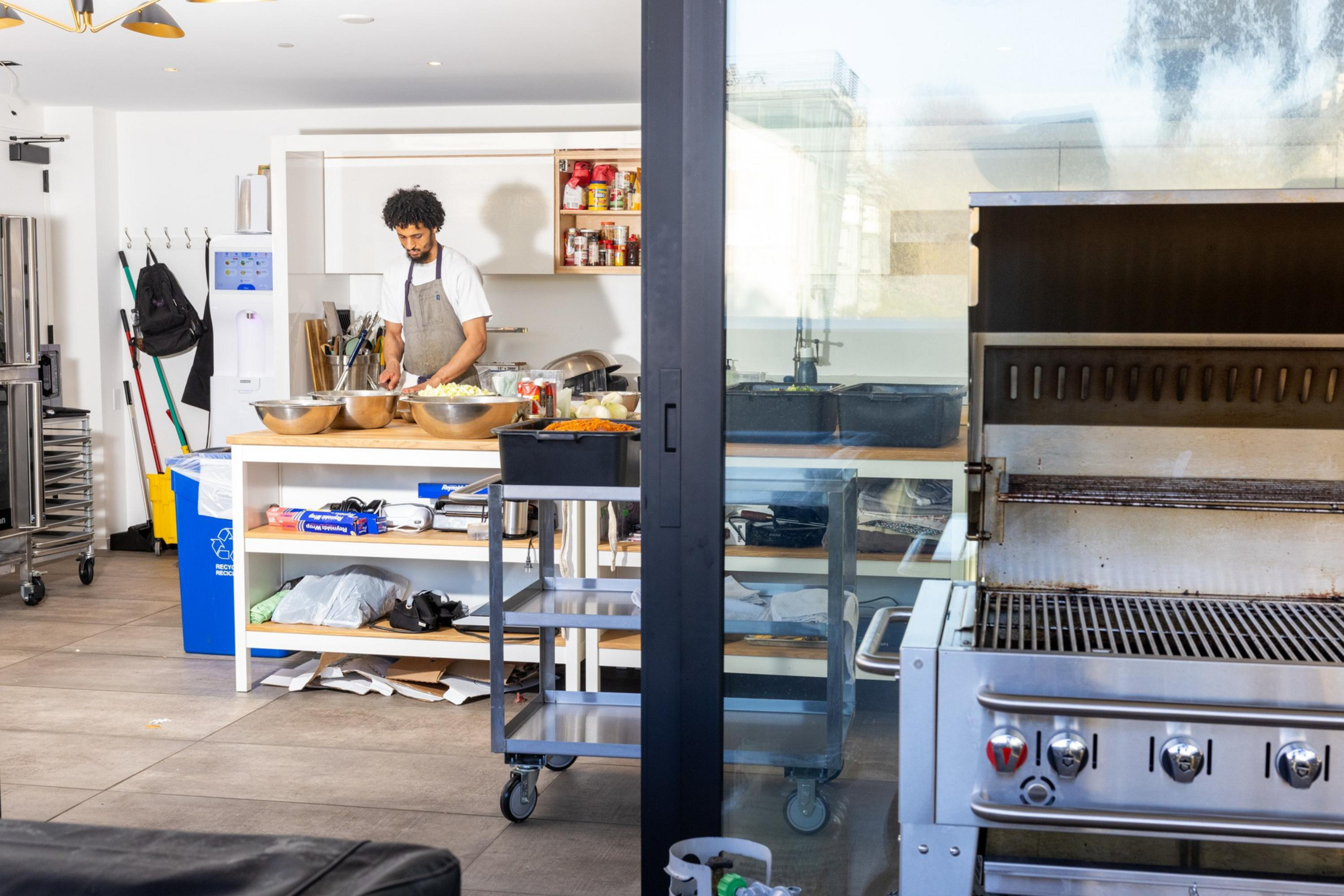 A man wearing an apron is preparing food at a kitchen counter with metal bowls, shelves, and cooking equipment around in a bright kitchen.