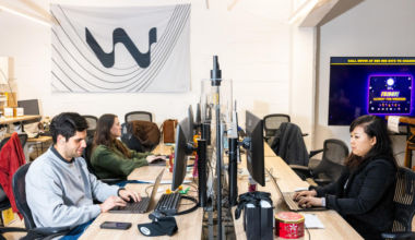 Several young adults work at computers in an office with an American flag on the wall, desks cluttered with water bottles, snacks, and electronics.