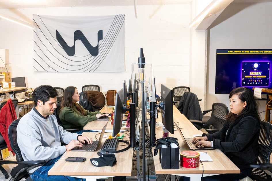 Several young adults work at computers in an office with an American flag on the wall, desks cluttered with water bottles, snacks, and electronics.