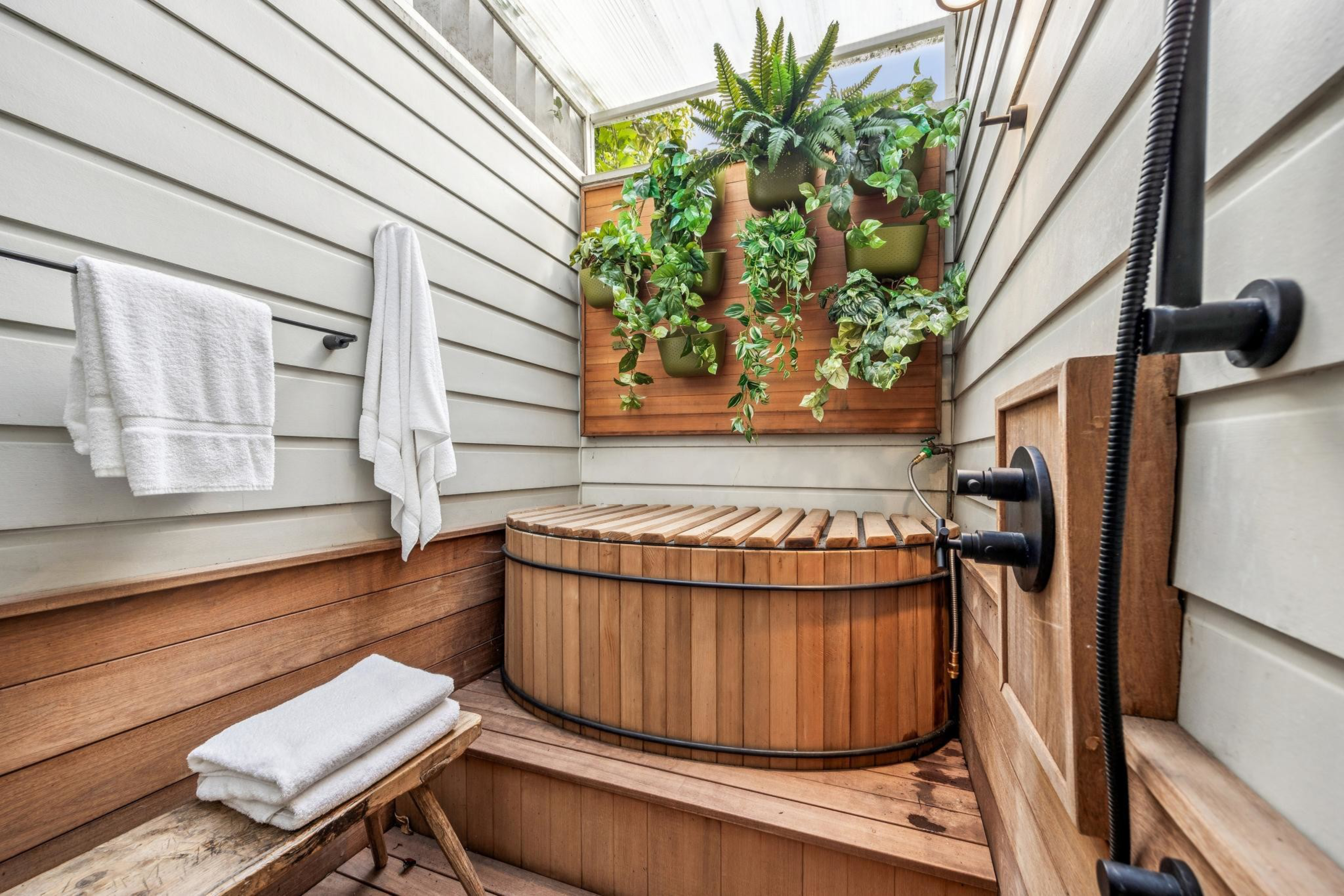 A small wooden hot tub sits in a narrow room with white-paneled walls, a wooden bench with folded towels, hanging white towels, a shower fixture, and green plants above.