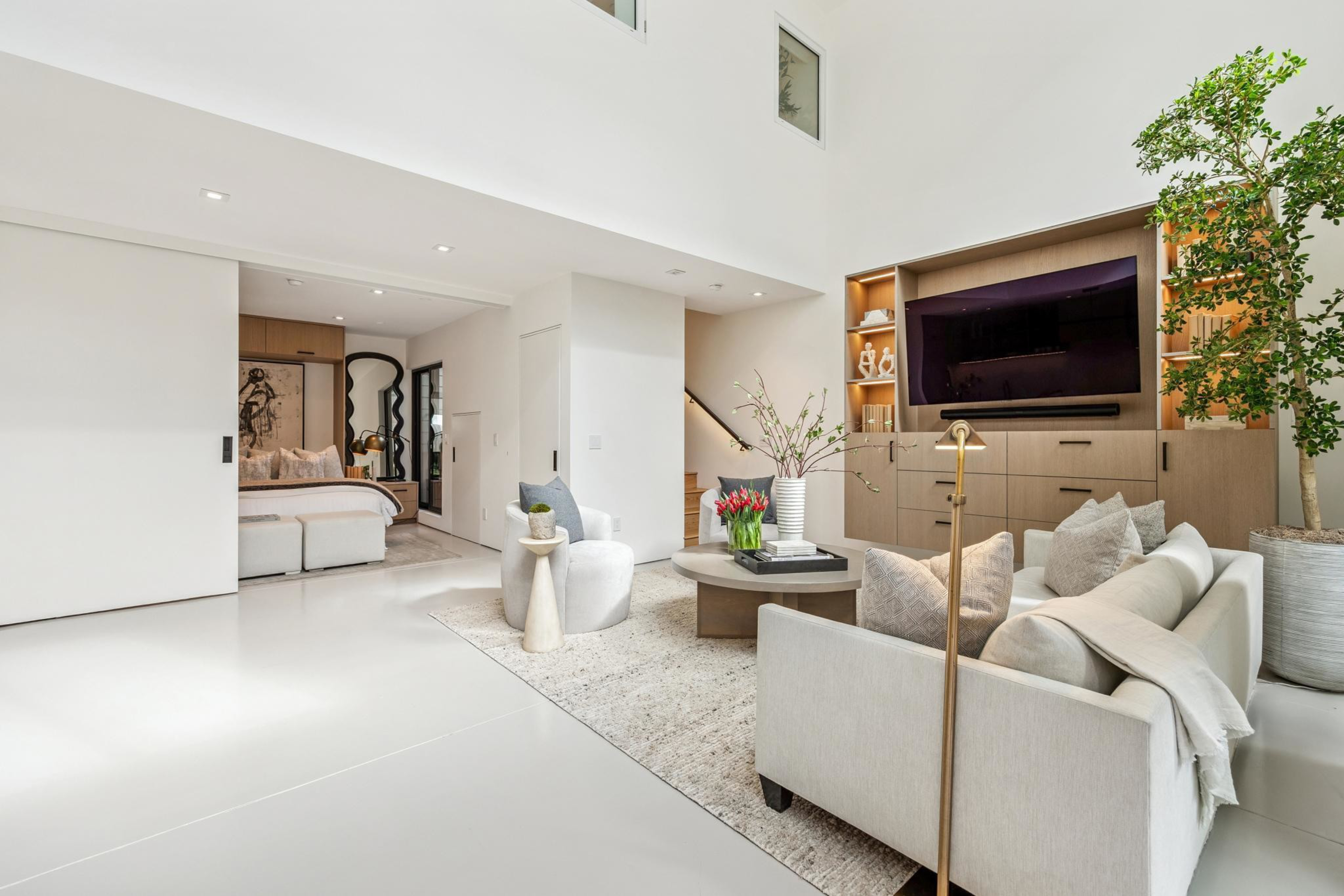 A bright, modern living room with beige furniture, a round coffee table, built-in wooden shelves, a large TV, and a potted tree near the corner.