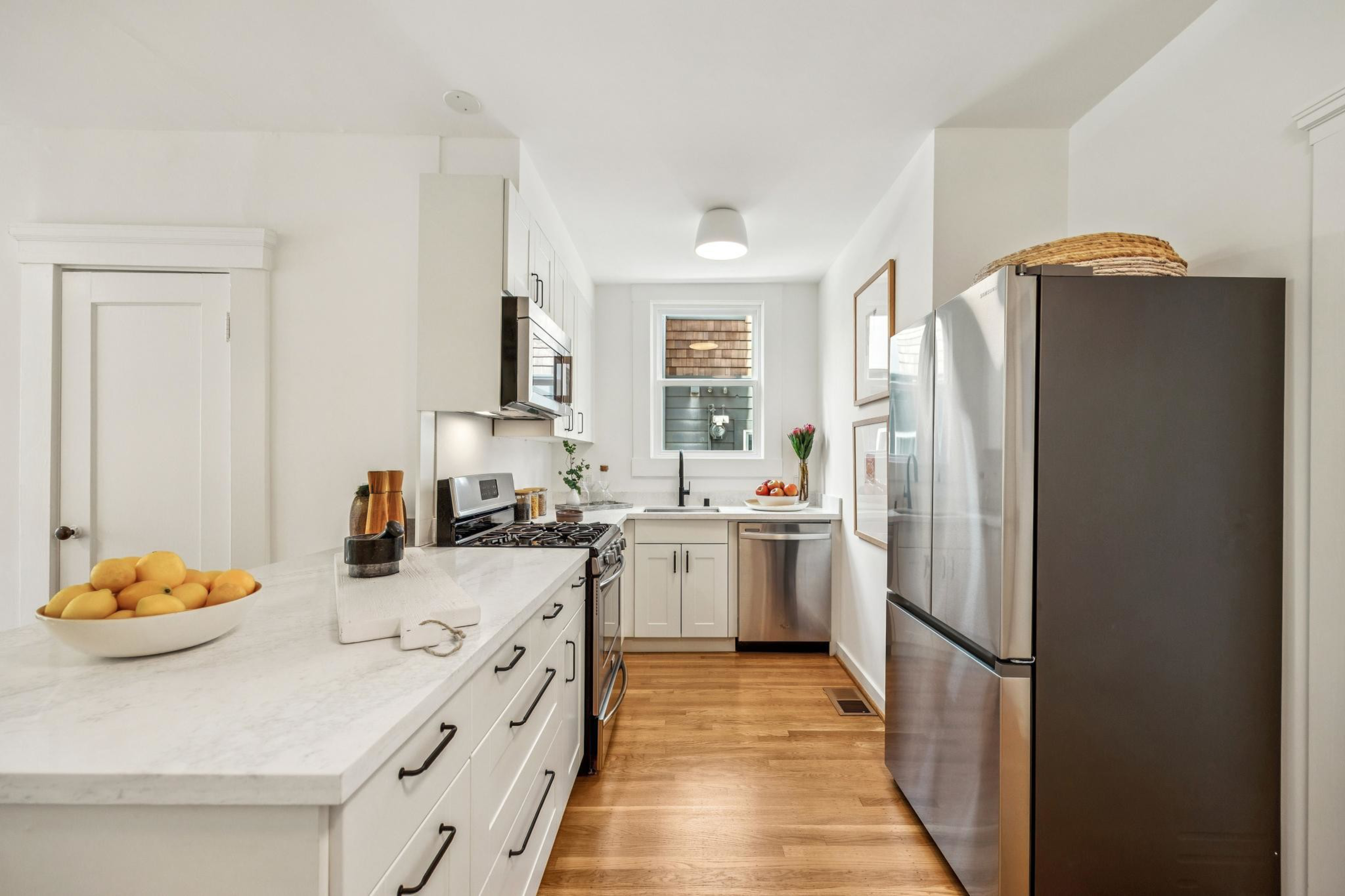 White kitchen with marble countertops, stainless steel appliances, wooden floor, bowl of lemons, and a window above the sink.