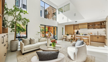 A bright, modern living room with tall windows, beige sofa, two round chairs, a round coffee table with flowers, and an open kitchen with wooden cabinets.