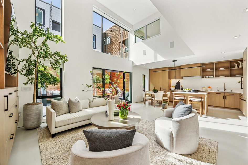 A bright, modern living room with tall windows, beige sofa, two round chairs, a round coffee table with flowers, and an open kitchen with wooden cabinets.