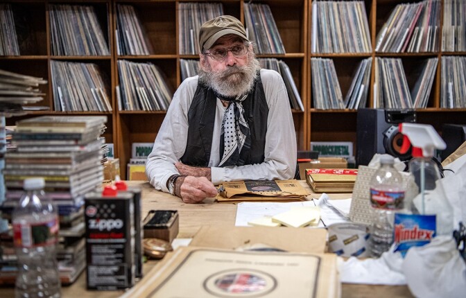 A man wearing an olive green baseball cap and wire rimmed eyeglasses leans on a table. The man has a white beard and is wearing a black and white polka dot scarf around his neck, a white long sleeved shirt and a black vest. On the table are stacks of CD's, cleaning supplies and a plastic water bottle. Behind him are shelves of records.