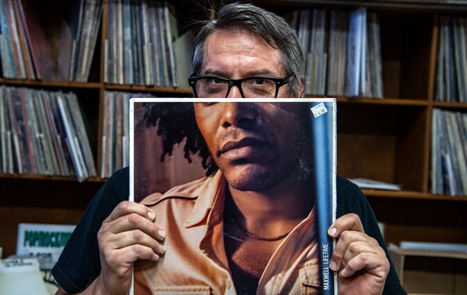 A man with salt and pepper hair, wearing black rimmed eyeglasses, holds up a record album with the lower half of a man's face.