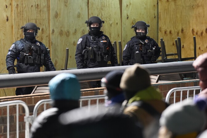 Three police officers in black riot gear stand behind a brick wall and stack chairs separated between a metal gate from people, who are out of focus in the foreground.