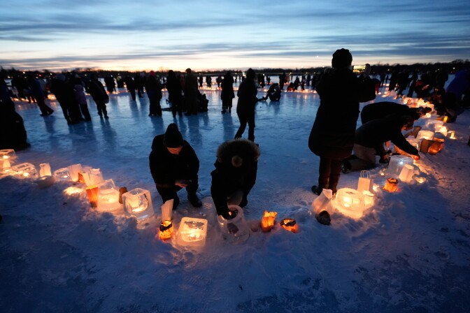 People set down candles and other items as others stand around in smaller groups on a frozen lake at sunset.