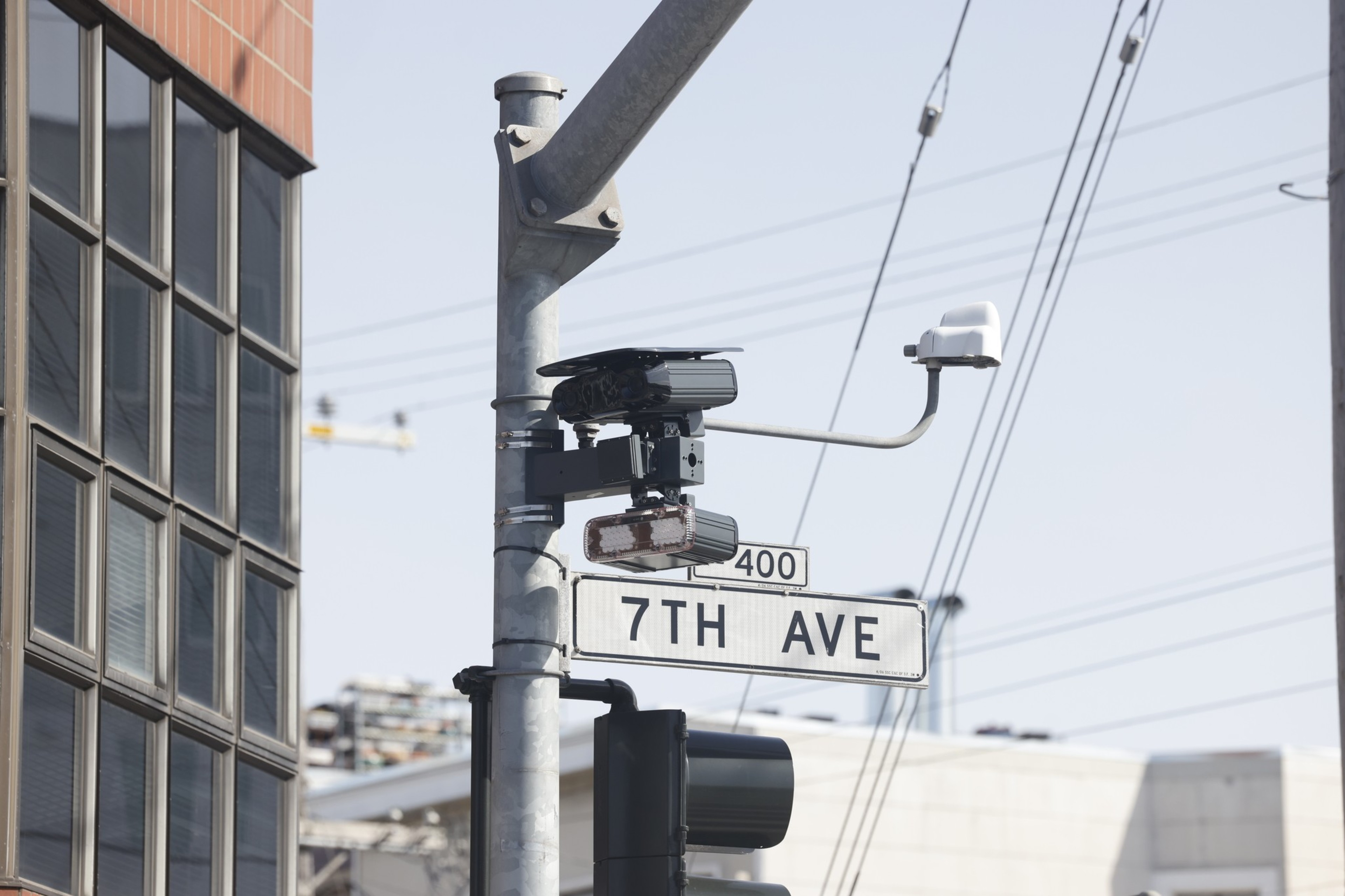 A street sign labeled “7TH AVE” is mounted on a pole with traffic cameras and a traffic light nearby, against a background of buildings and power lines.