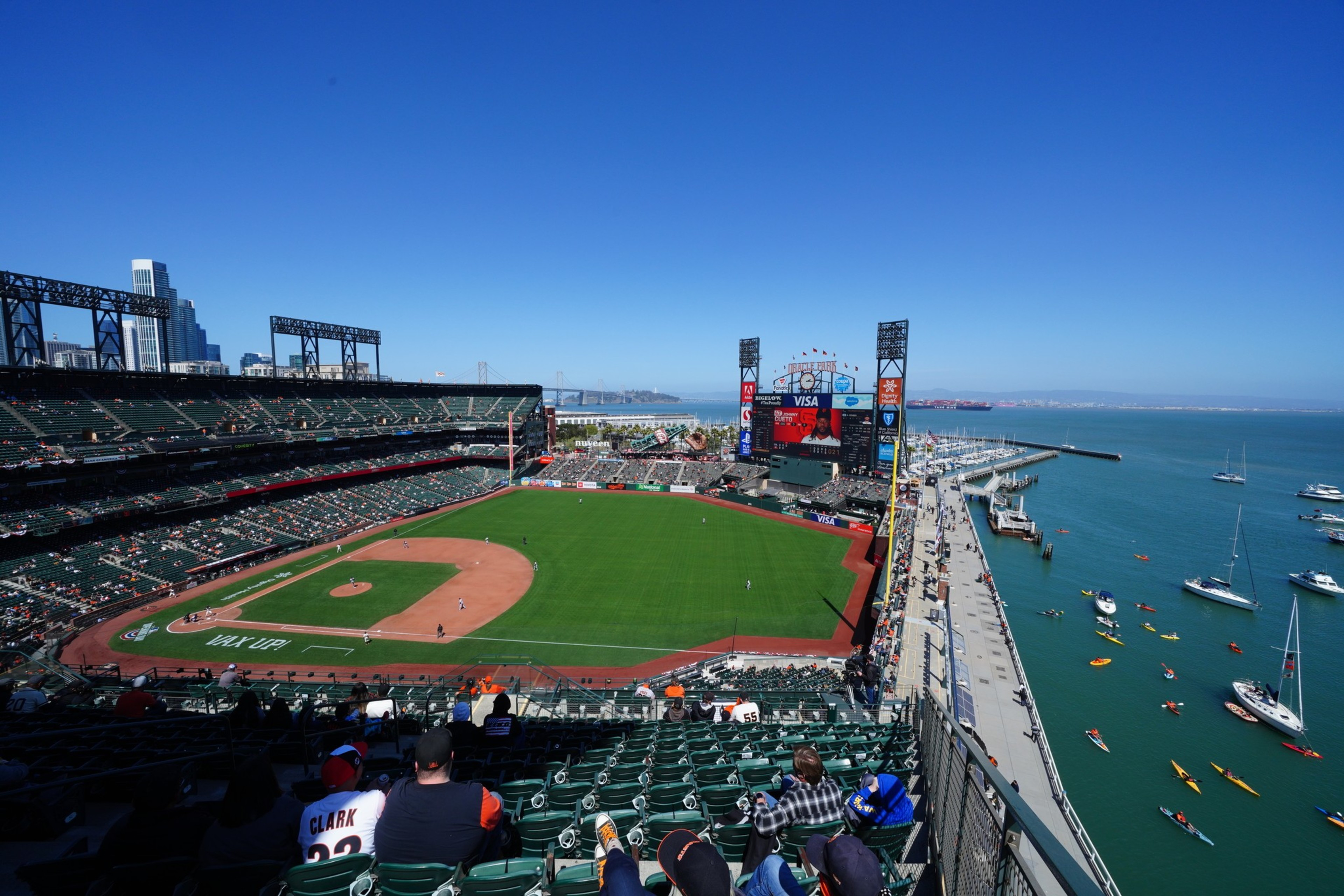 A sunny baseball stadium overlooks a green field with players, a dock with boats and kayaks, and a city skyline in the background under a clear blue sky.
