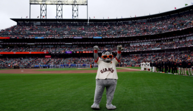 A mascot in a Giants jersey with orange glasses stands on a baseball field with arms raised, while players line up and spectators fill the stadium seats.
