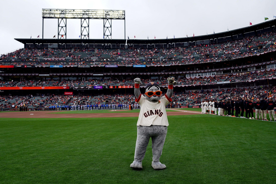 A mascot in a Giants jersey with orange glasses stands on a baseball field with arms raised, while players line up and spectators fill the stadium seats.