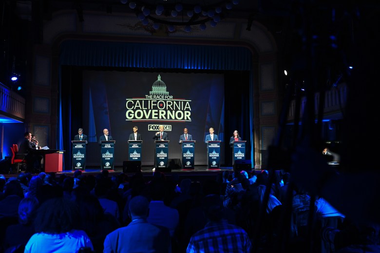Candidates stand at podiums on stage during a televised debate titled “The Race for California Governor,” while moderators sit at a desk to the side and an audience watches from the theater seating.