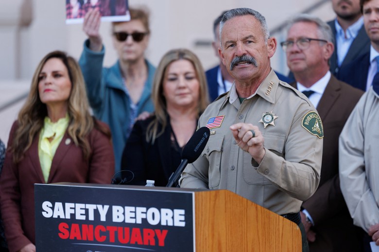A person with a mustache and a sheriff’s uniform speaks at a podium, saying, “Safety Over Sanctuary.” In the background is a row of people dressed professionally and listening to the speech.