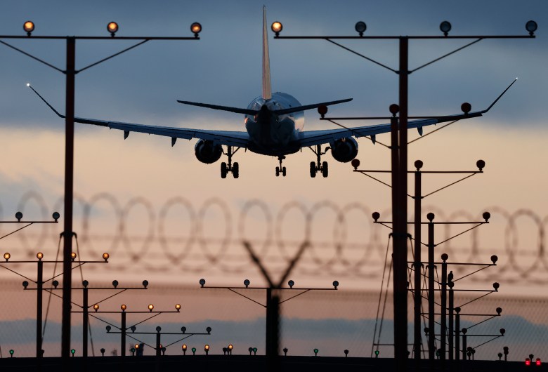 The back of a plane taking off from an airport is seen between runway landing lights, with an out-of-focus fence with barbed wire in the foreground.