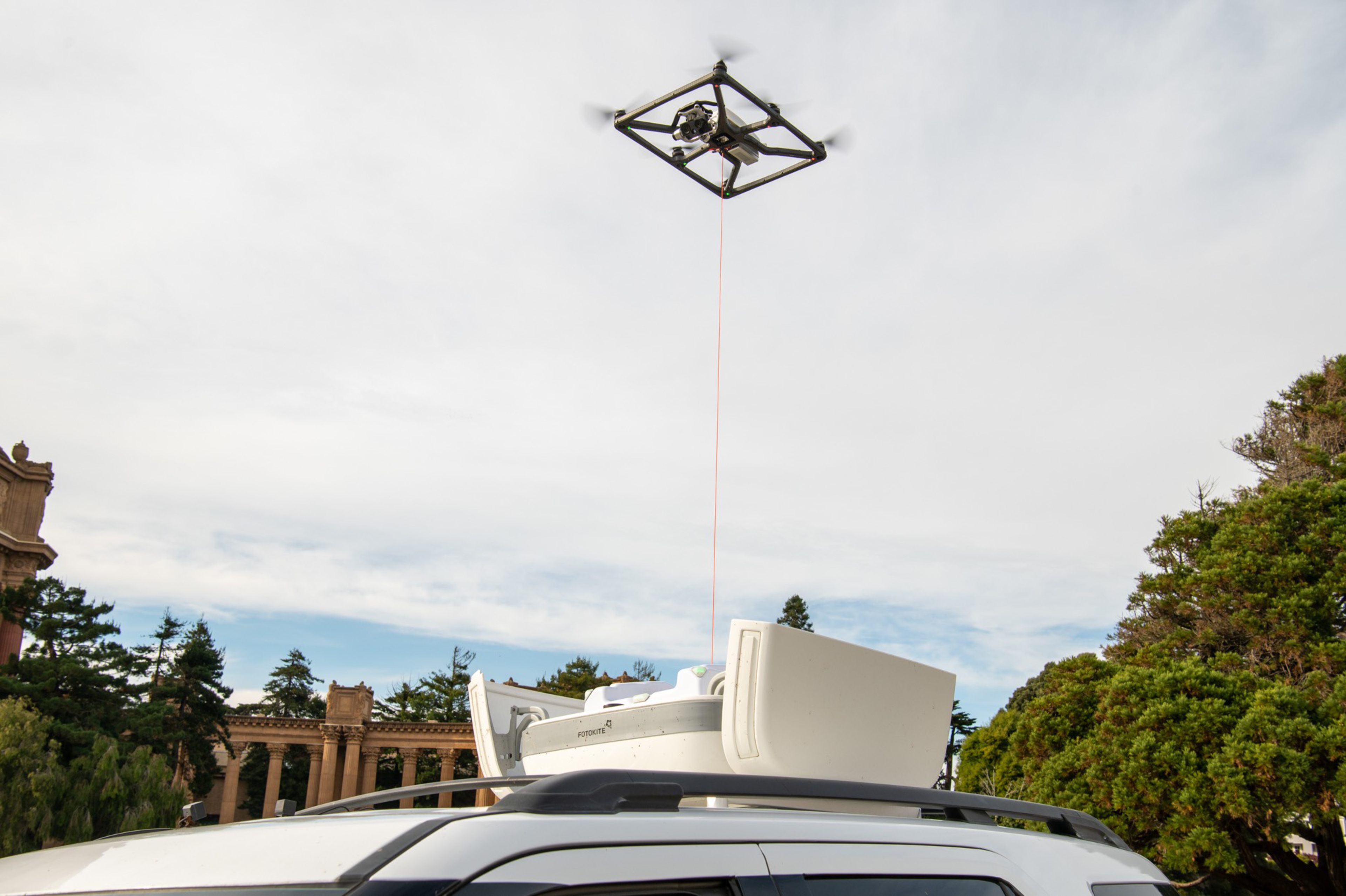 A square-shaped drone hovers above a white car, lifting a sealed package attached by a red cable on a clear, partly cloudy day.