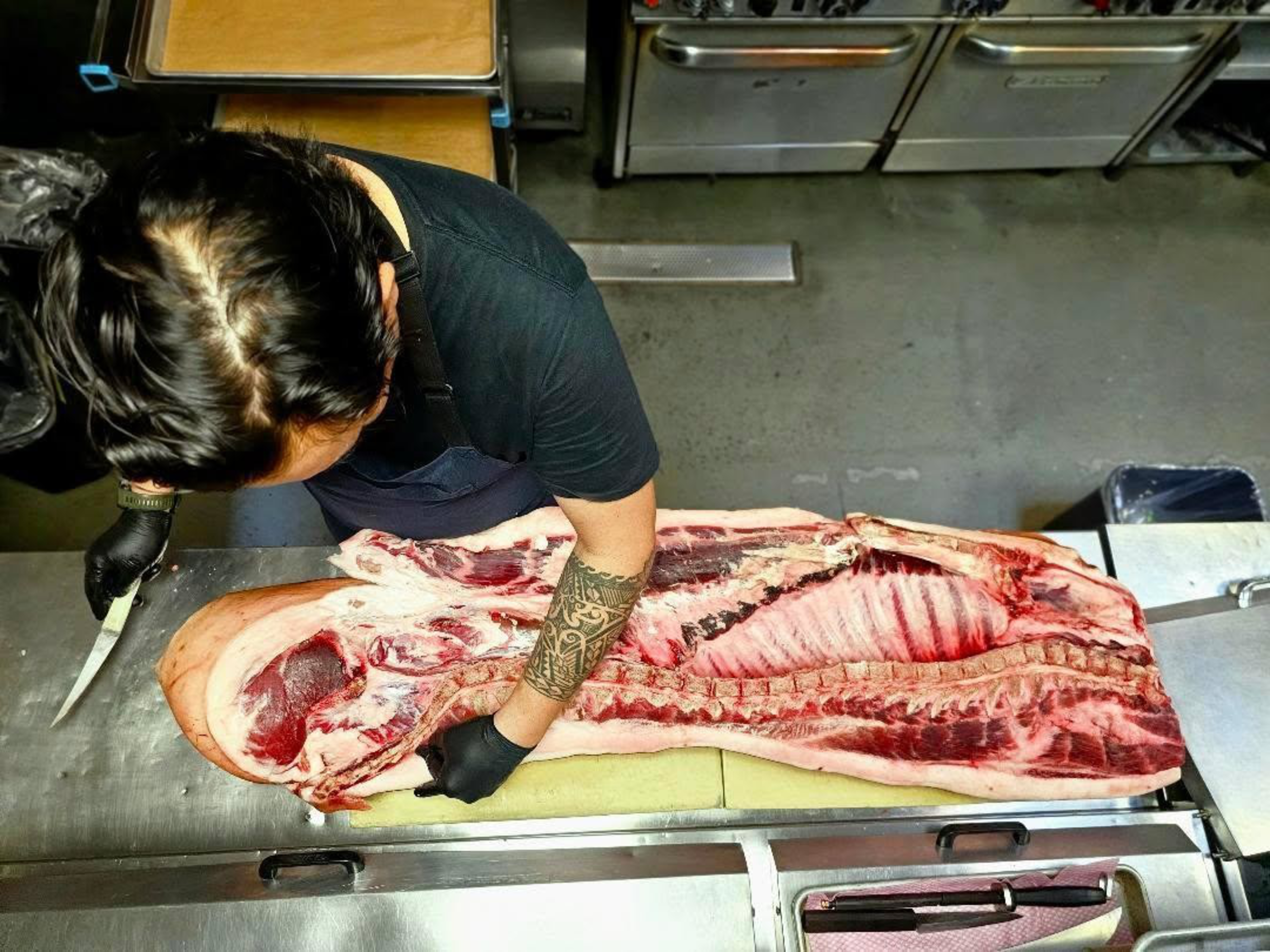 A person with a tattooed arm and black gloves is cutting a large, split open pig carcass on a metal table in a kitchen setting.