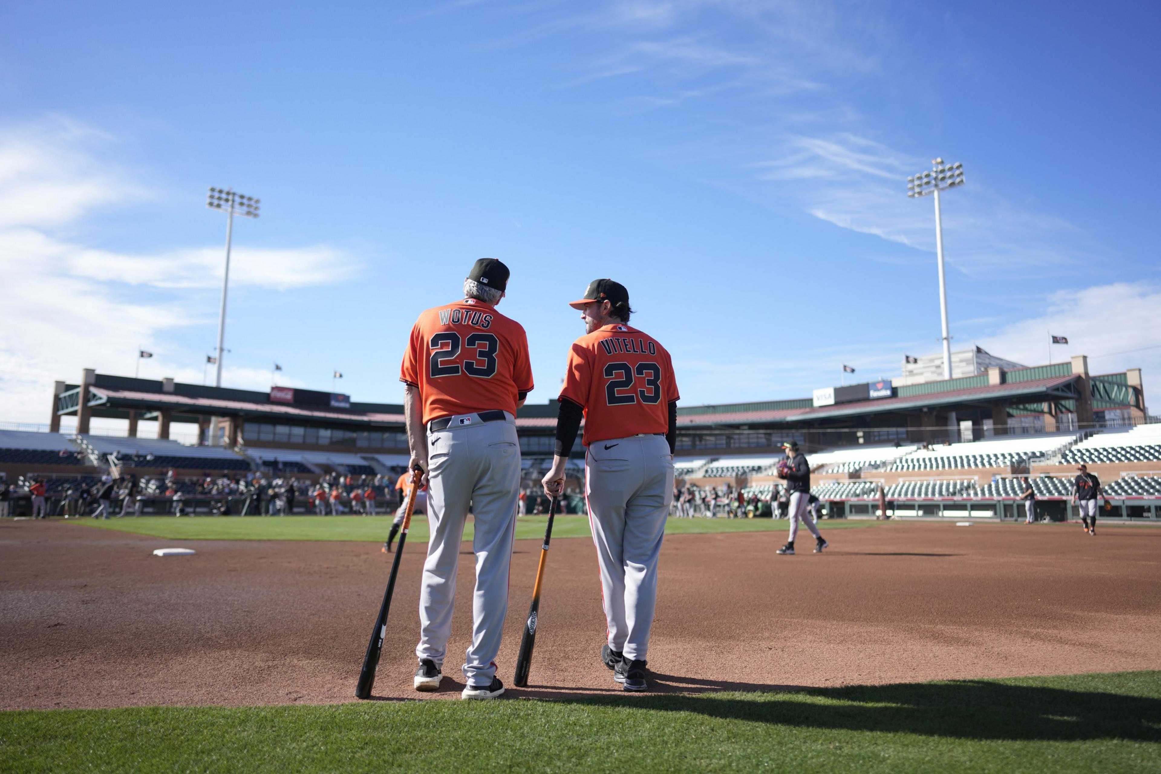 Two baseball players in orange jerseys with number 23 stand side by side on the field, holding bats and facing away toward the stadium.