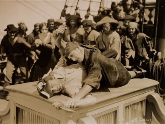 A black-and-white film still of a light-skinned man holding down another man with a knife in his hand while a group of pirates looks on. 