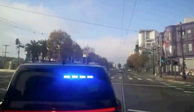 A dark police SUV with flashing blue lights is stopped at an urban intersection with palm trees, buildings, and a green traffic light ahead.