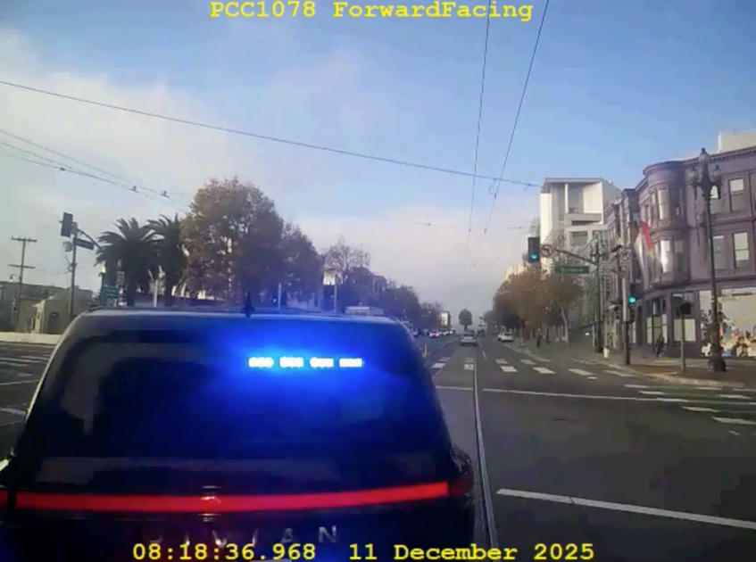 A dark police SUV with flashing blue lights is stopped at an urban intersection with palm trees, buildings, and a green traffic light ahead.