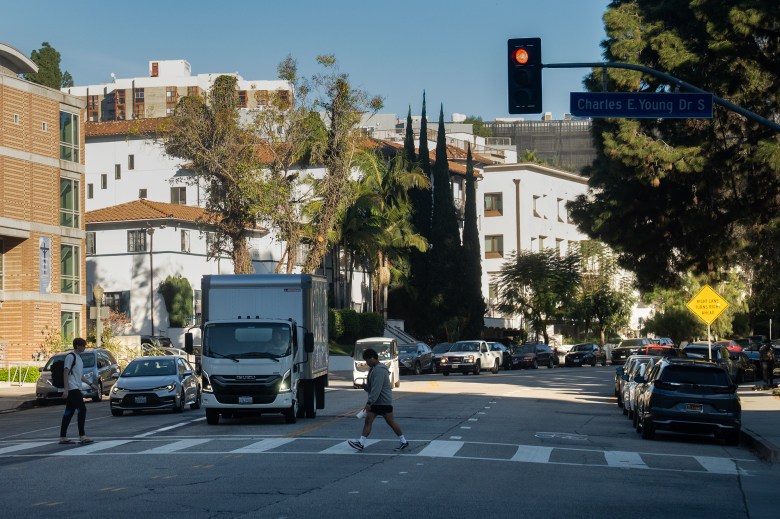 People use a cross walk along a street intersection as a white semi-truck and a gray car sit idle.