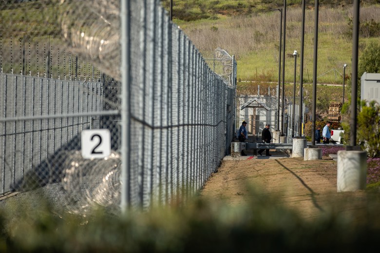 A large detention facility complex sits behind tall chain-link fencing topped with coils of razor wire.