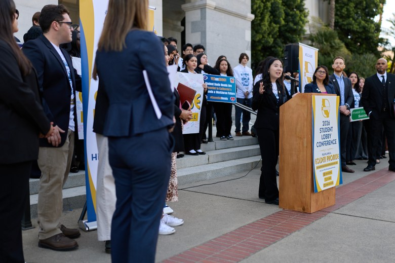 A person wearing a black suit stands in front of a podium as students and supporters stand behind them during a press conference.