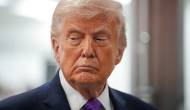 A man with light blond hair, wearing a dark suit, white shirt, and purple patterned tie, looks sideways with a serious expression.