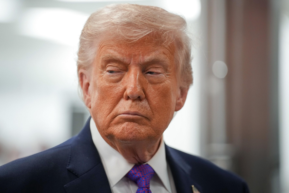 A man with light blond hair, wearing a dark suit, white shirt, and purple patterned tie, looks sideways with a serious expression.