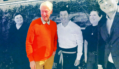 Five people stand outside against a brick wall with ivy; one man wears a bright orange sweater, others are dressed in black, white, and gray.