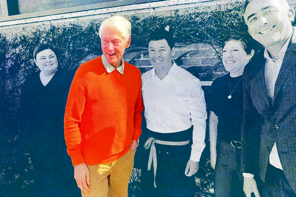 Five people stand outside against a brick wall with ivy; one man wears a bright orange sweater, others are dressed in black, white, and gray.