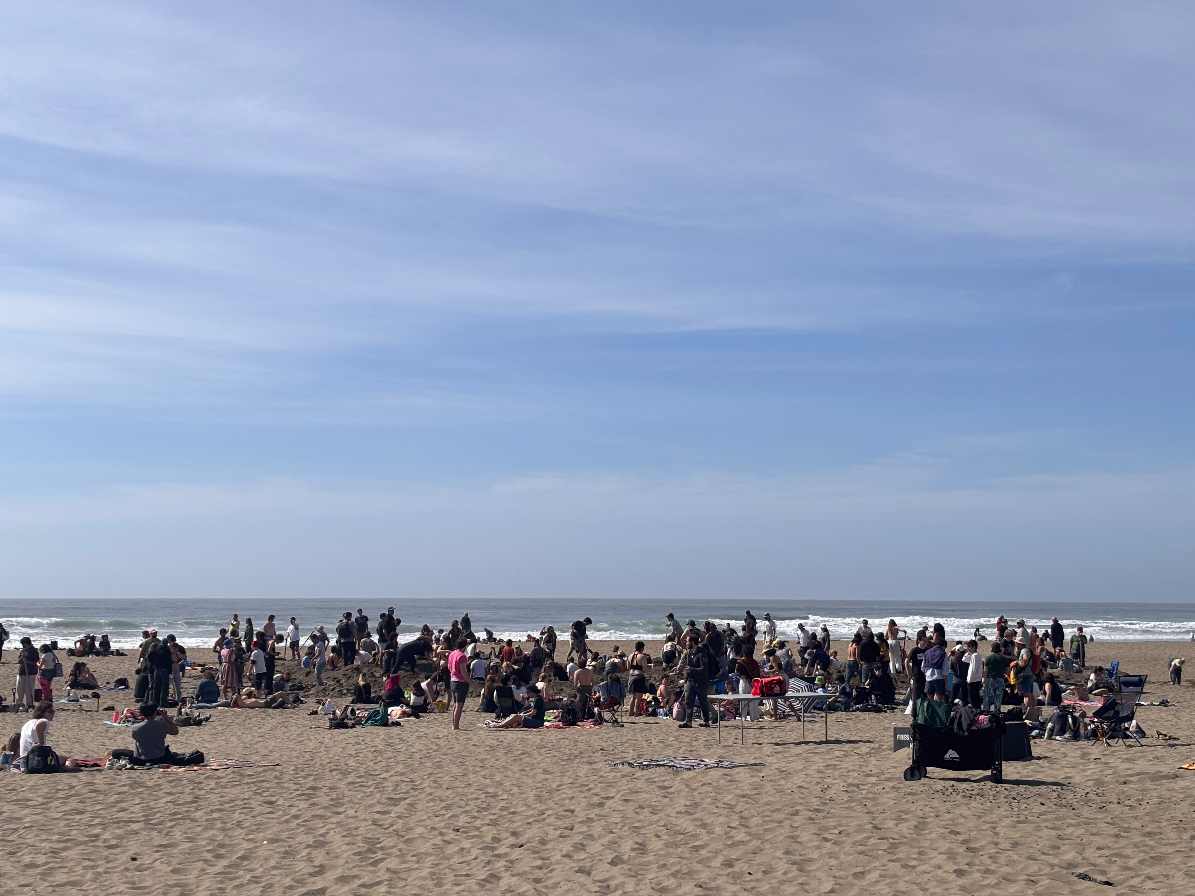 A large group of people gathers on a sandy beach under a blue sky, with the ocean waves visible in the background.