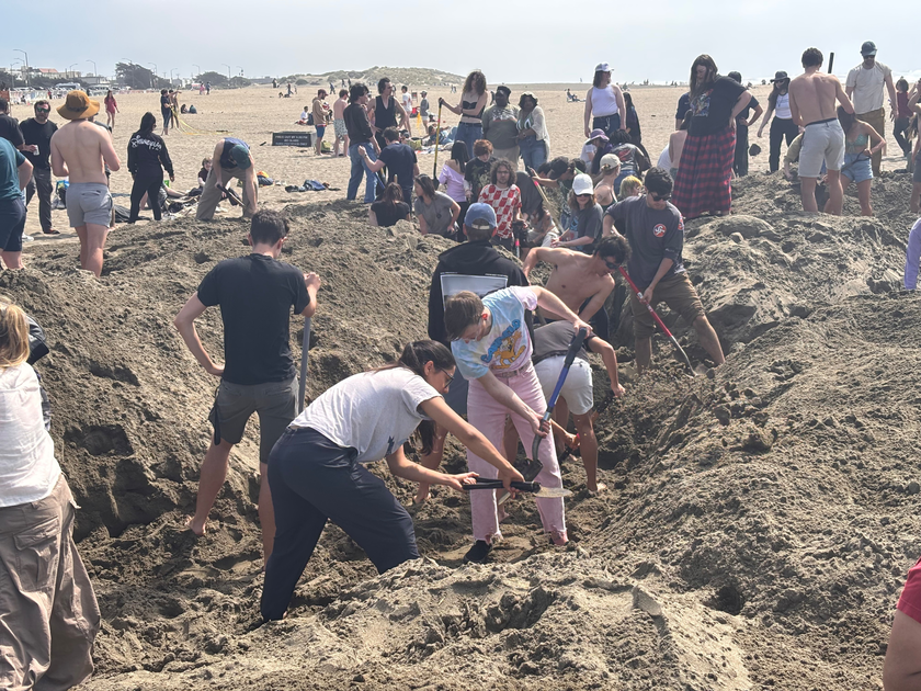 Many people on a sandy beach are digging a large hole together using shovels and other tools on a sunny day.