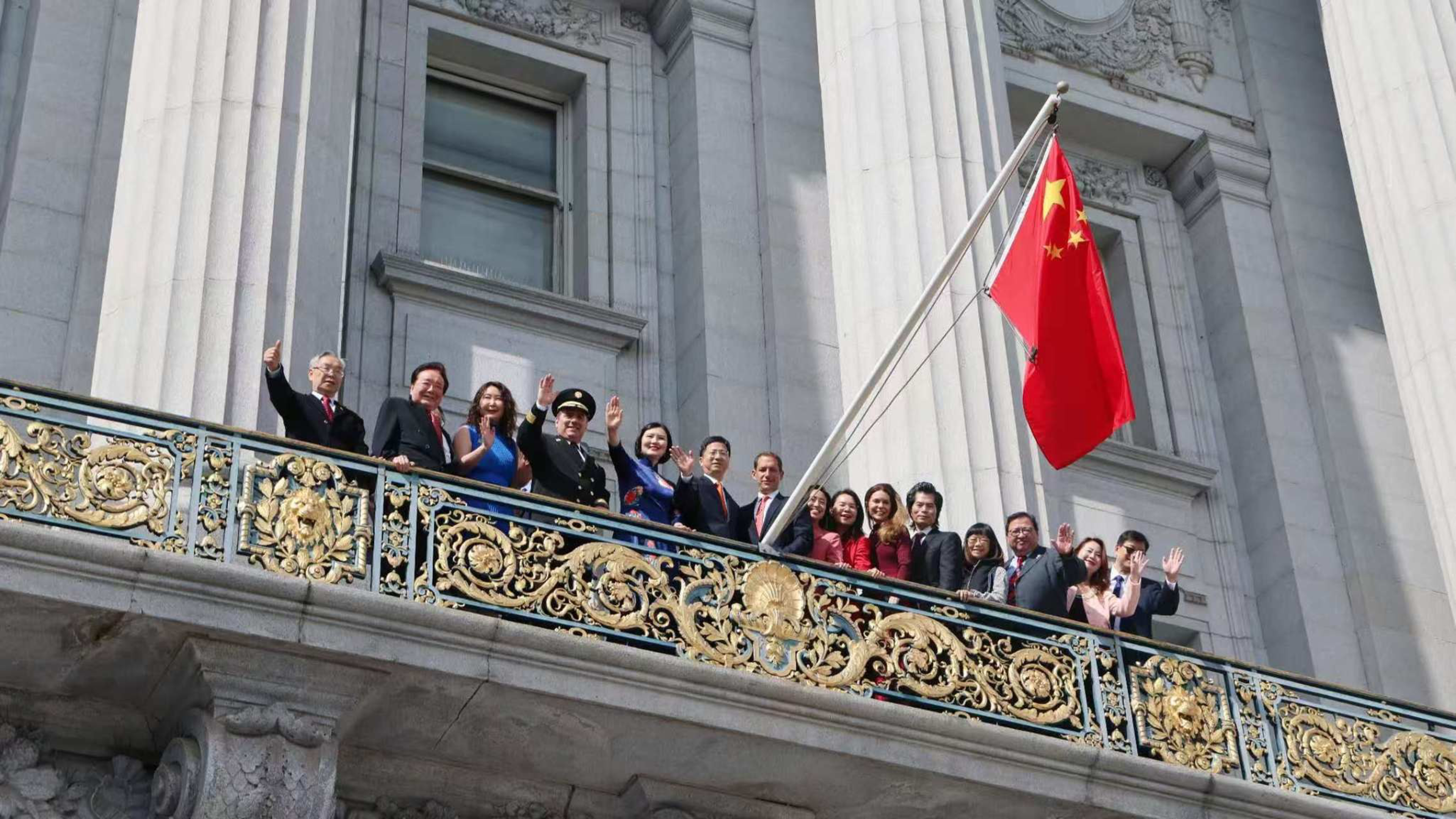 A group of sixteen people in formal attire stand on a balcony with ornate gold railings, waving beneath a large Chinese flag.