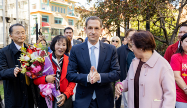 A man in a suit walks surrounded by smiling people; one woman holds a bouquet, and the group seems happy and engaged in conversation.