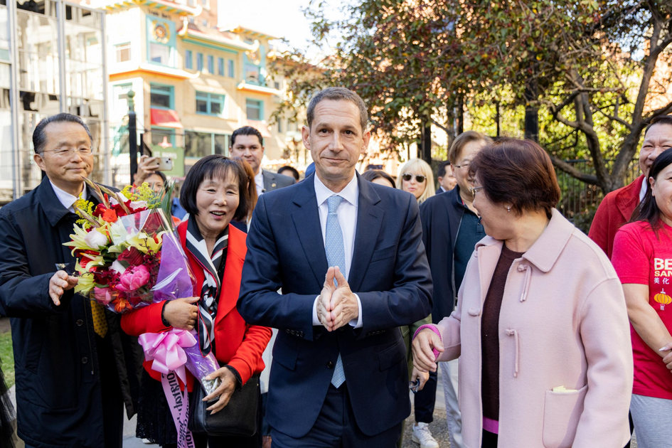 A man in a suit walks surrounded by smiling people; one woman holds a bouquet, and the group seems happy and engaged in conversation.