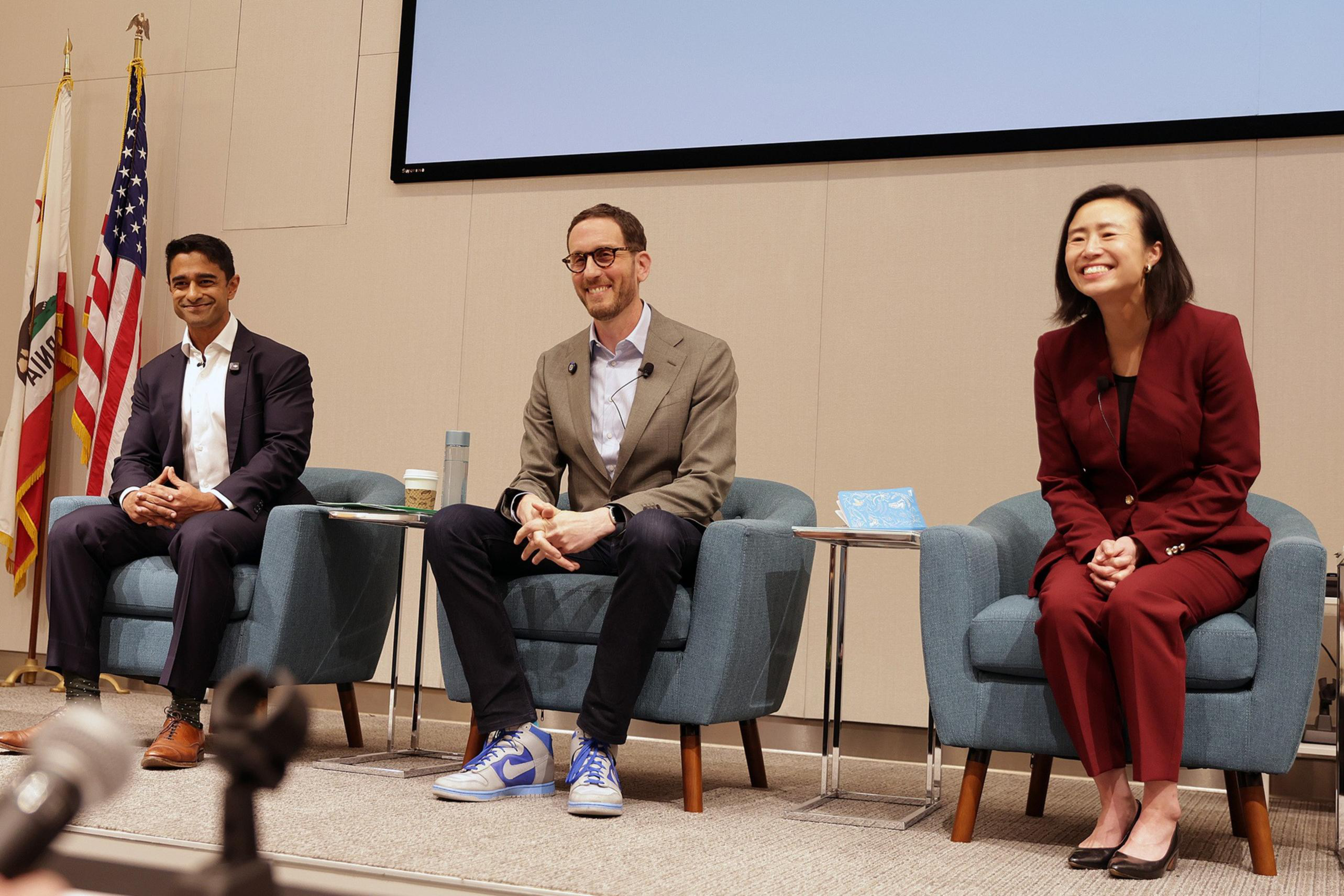 Three people, two men and one woman, sit on armchairs on a stage, smiling, with American and California flags in the background.