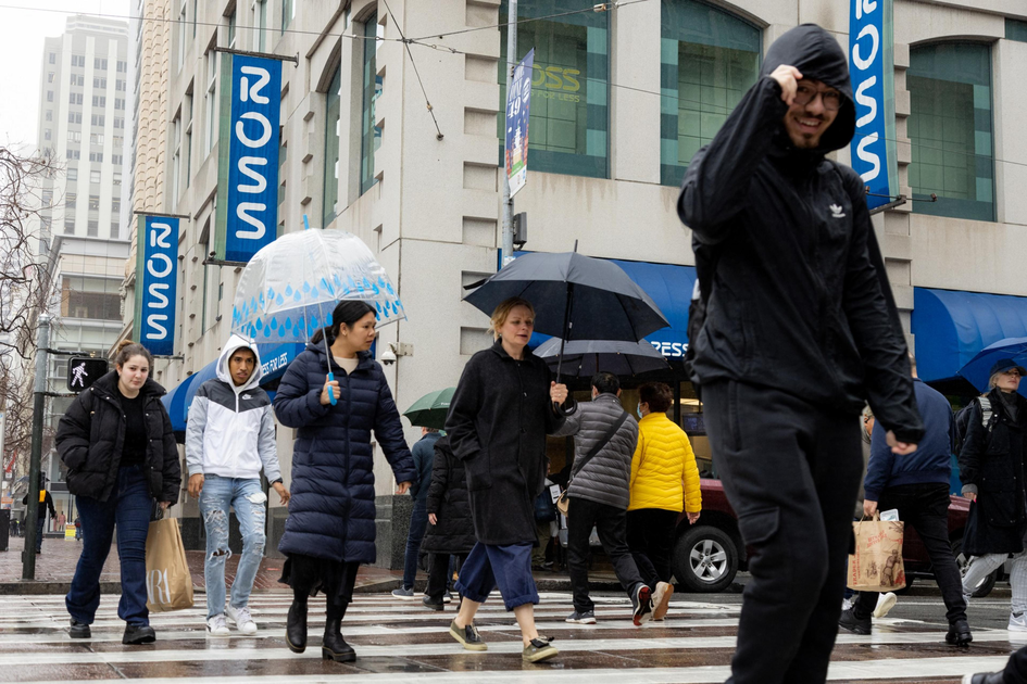 People wearing coats and carrying umbrellas walk across a city street on a rainy day near a store with blue "ROSS" signs.