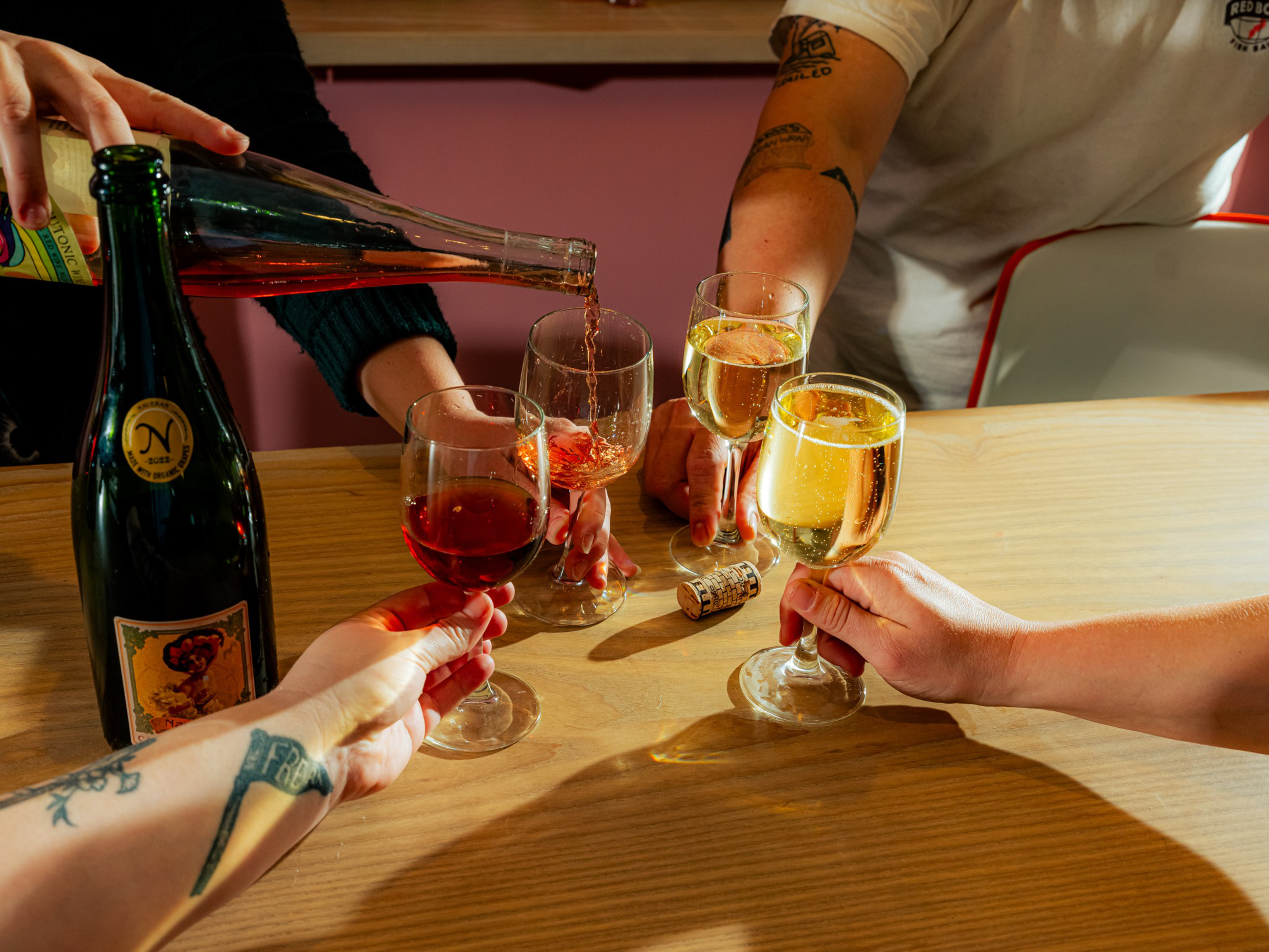 Four people are holding glasses filled with red and white sparkling wine, one glass is being filled from a bottle on a wooden table.