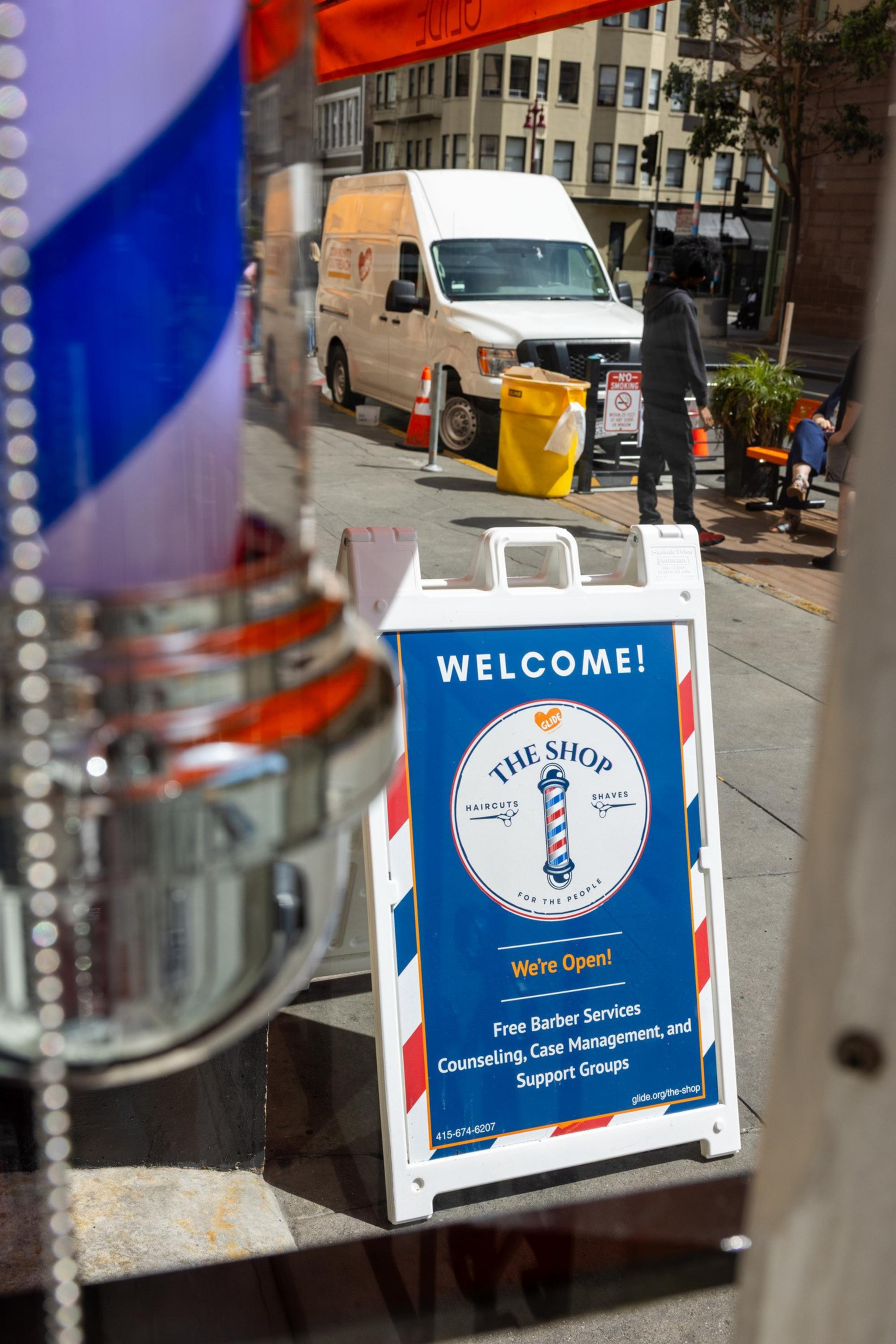 A sidewalk sign welcomes people to The Shop, offering free barber services, counseling, case management, and support groups, with a white van and people in the background.