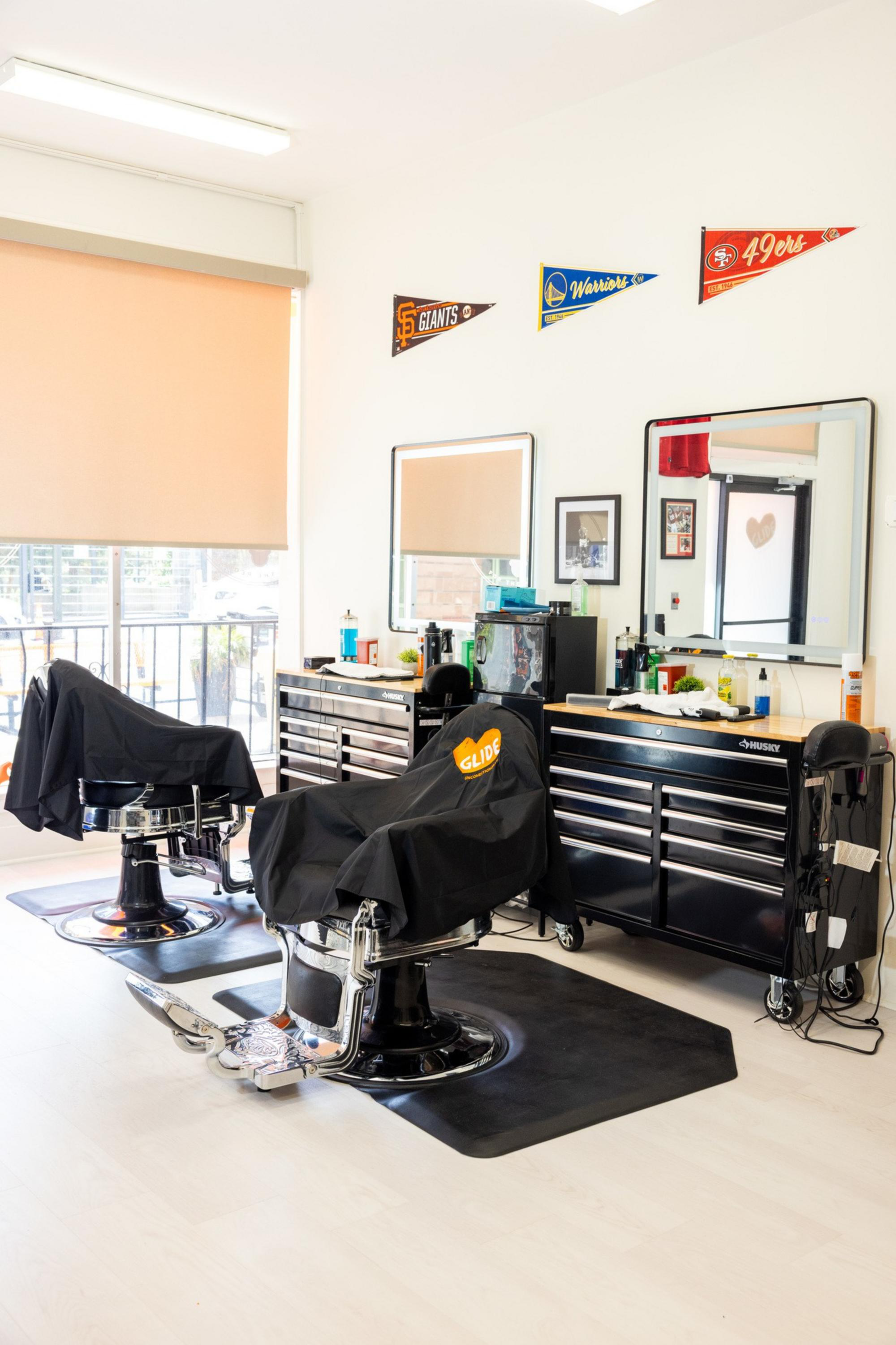 Two black barber chairs with covers face mirrors above black tool cabinets in a bright room decorated with sports pennants on the wall.