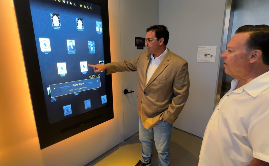 Navy SEAL Museum Executive Director Brian "Beef" Drechsler is shown demonstrating the wall of remembrance display to KPBS reporter John Carroll on March 23, 2026.